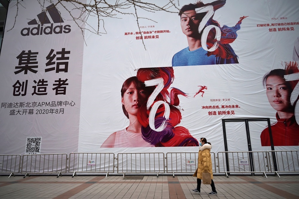 Mulher com máscara de proteção contra coronavírus passa por banner da Adidas 