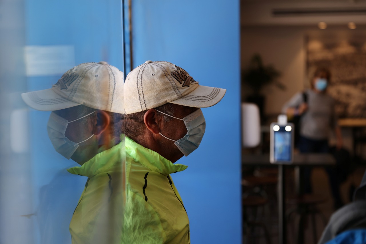 Homem usando máscara é refletido no vidro ao entrar em espabelecimento de Israel