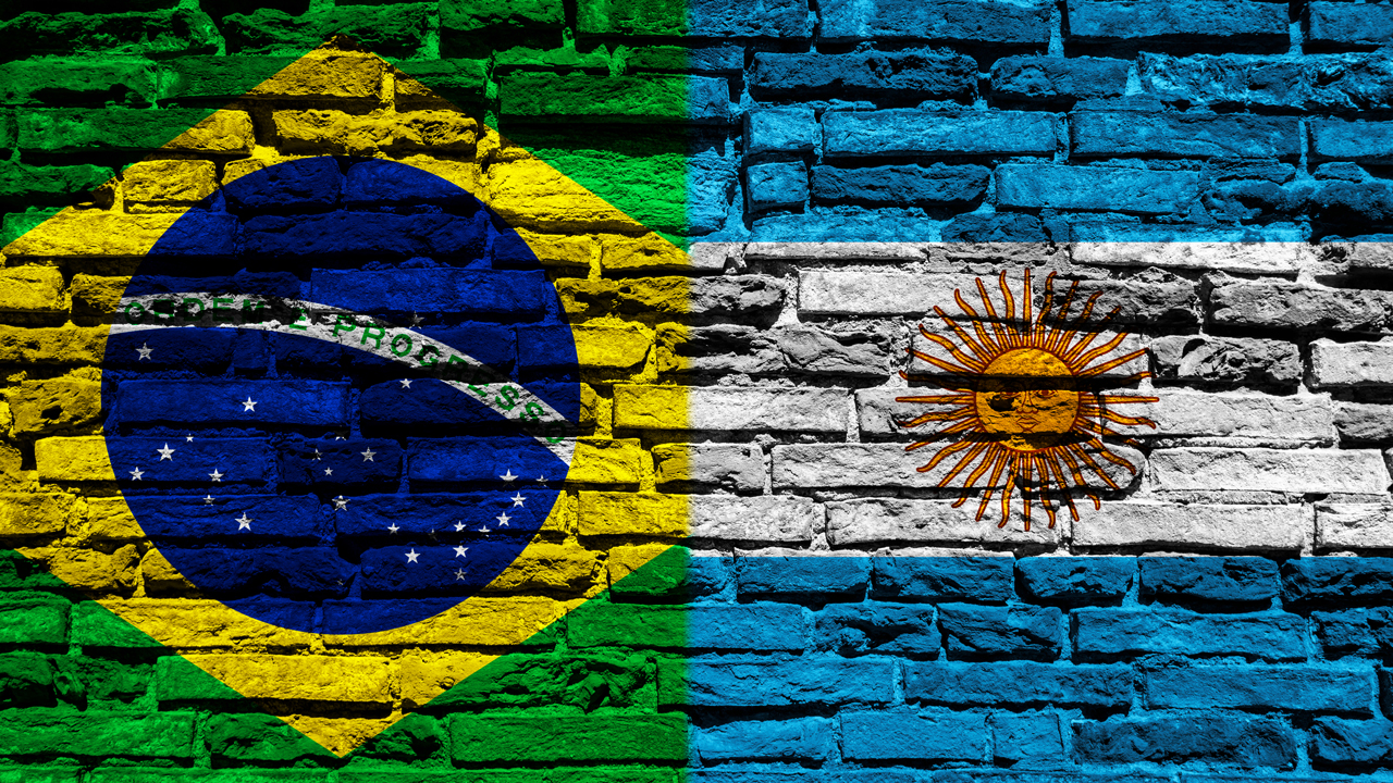Bandeiras do Brasil e Argentina lado a lado pintadas em um muro de tijolos. 