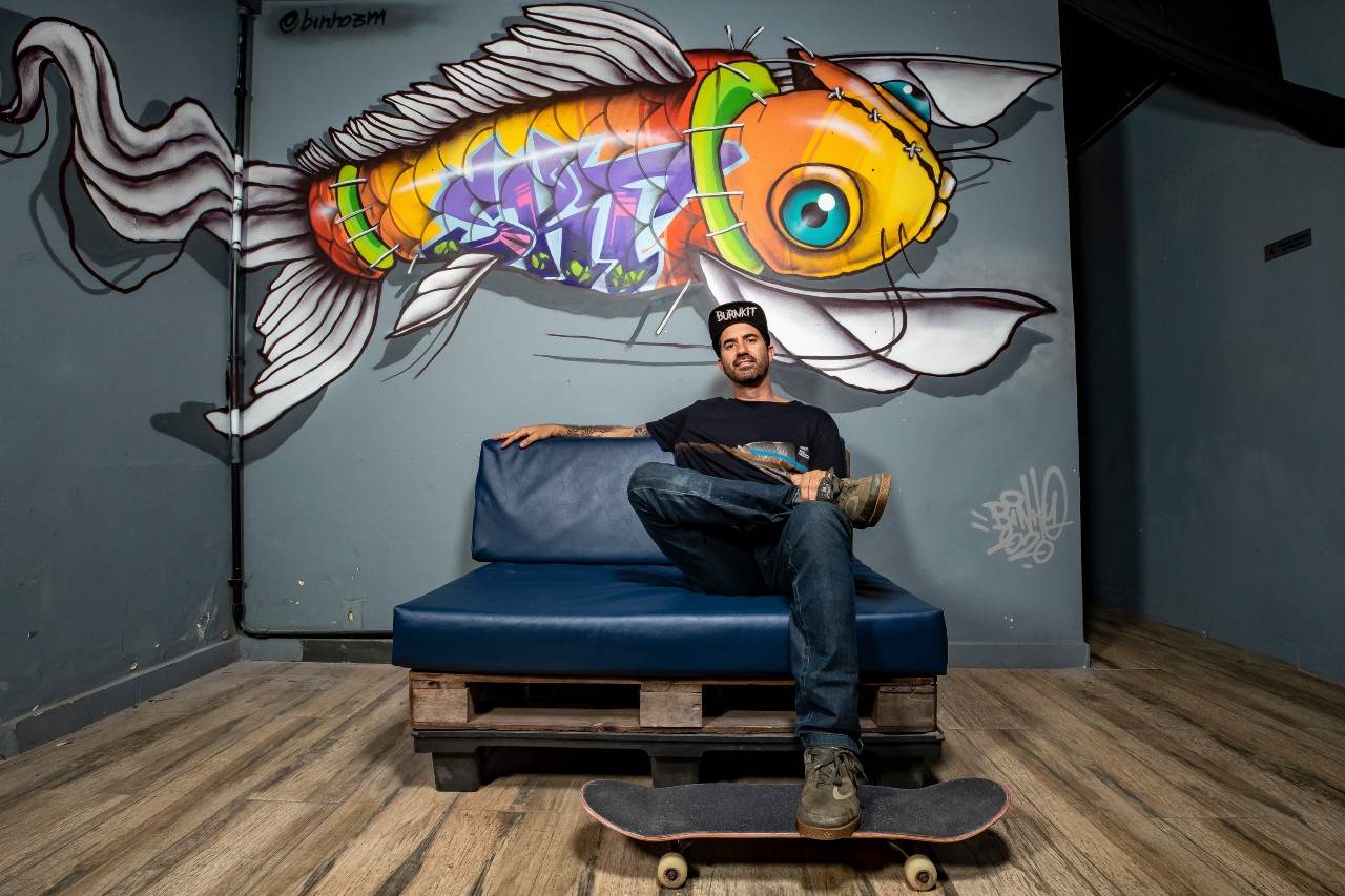 Bob Burnquist sentado em um sofá em frente a um grafite com o skate no pé Bob Burnquist sentado em um sofá em frente a um grafite com o skate no pé