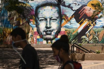 Caracas, Venezuela: Homem e mulher de máscaras andam em frente à parede com a imagem do falecido presidente Hugo Chávez