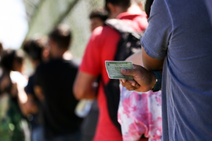 A imagem mostra uma fila de pessoas. Seus rostos estão desfocados e mais próxima segura um título de eleitor.