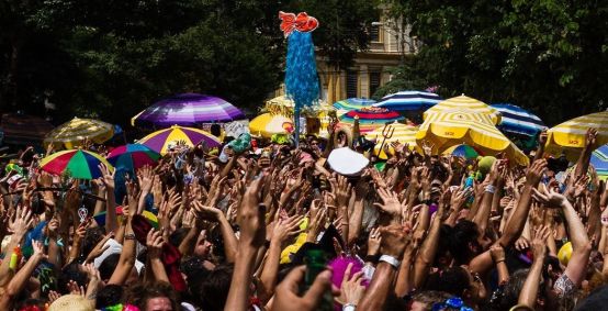 Pessoas aglomeradas em um bloco de carnaval em são paulo com as mãos para cima e guarda-sóis ao fundo