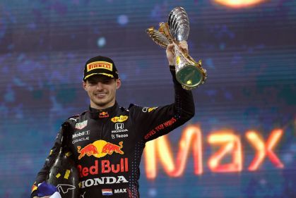 Foto de Max Verstappen, piloto de Fórmula 1 da Red Bull, segurando o prêmio do GP de Abu Dhabi. Ele está de macacão preto, boné e erguendo o troféu com a mão esquerda