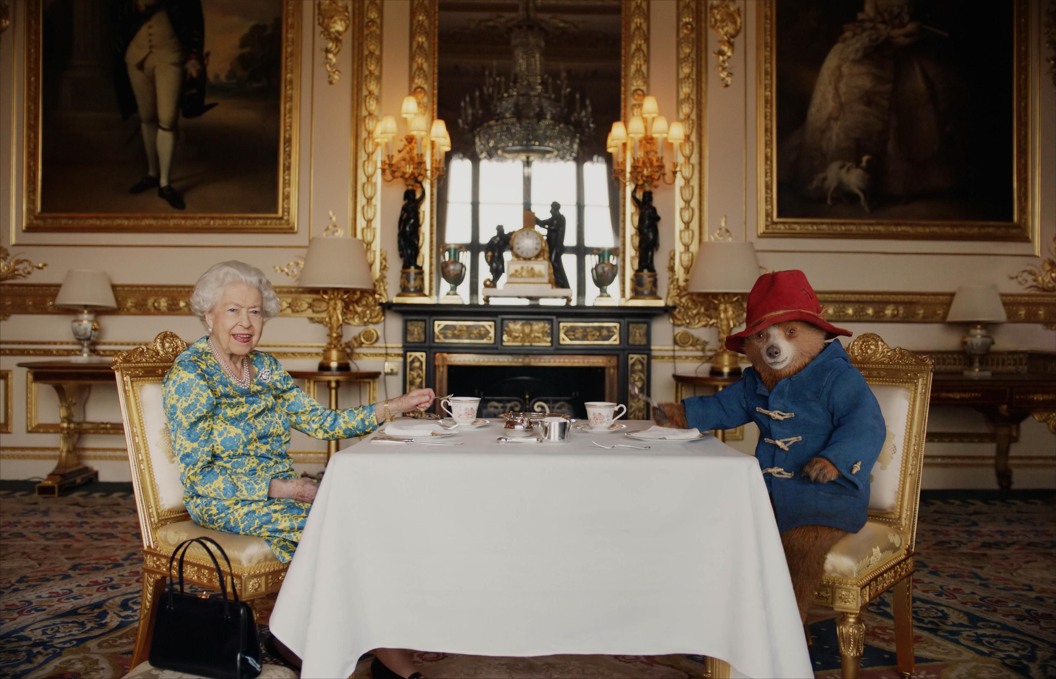 A rainha Elizabeth usou um vestido floral azul e amarelo para a hora do chá com ninguém menos que Paddington Bear, que compareceu com seu casaco azul e chapéu vermelho. A rainha revelou-se uma grande fã do lanche favorito de Paddington - sanduíches de marmelada - enquanto tirava um de sua bolsa preta/ Buckingham Palace/Studio Canal