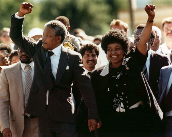 África do Sul, Cidade do Cabo, 11/02/1990. Cercado por uma multidão e de mãos dadas com a mulher Winnie, Nelson Mandela faz gesto com o punho fechado, sinal de oposição, após ser solto da prisão Victor Vester, na Cidade do Cabo. O líder sul-africano permaneceu preso por 27 anos • AP