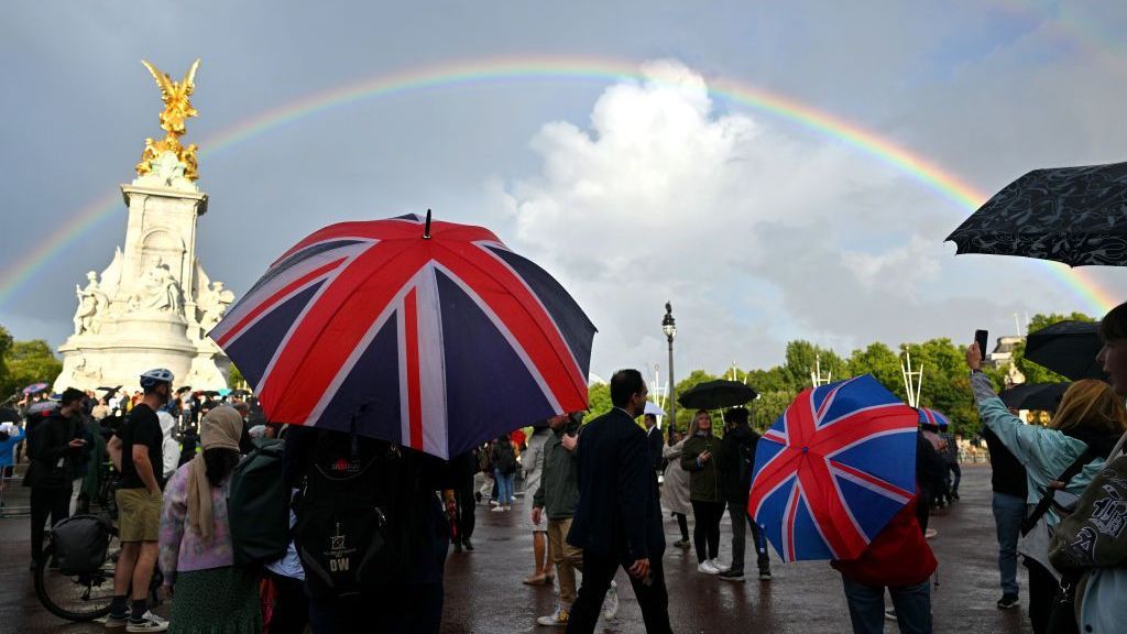 Um homem olha segurando um guarda-chuva da bandeira do Reino Unido enquanto um arco-íris é visto fora do Palácio de Buckingham em 08 de setembro de 2022 em Londres, Inglaterra
