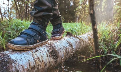 Close-up of child's feet in shoes crossing a forest stream along a tree trunk. The concept of solo walks with a pole in woodland