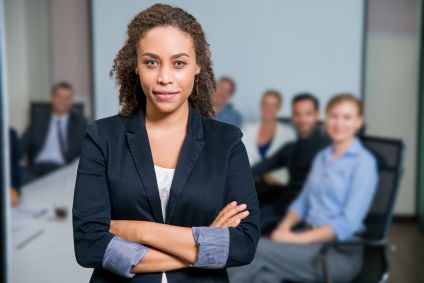 Portrait of female African American executive manager standing with crossed arms in meeting room, business people sitting at office desk on background