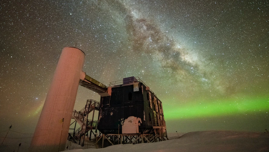 O detector IceCube é visto sob um céu noturno estrelado, com a Via Láctea aparecendo sobre auroras baixas ao fundo. / Yuya Makino/IceCube/NSF