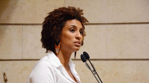 A ex-vereadora Marielle Franco discursa em frente a um microfone usando um blazer branco e brincos coloridos. Tem a pele negra, cabelo crespo e usa maquiagem.
