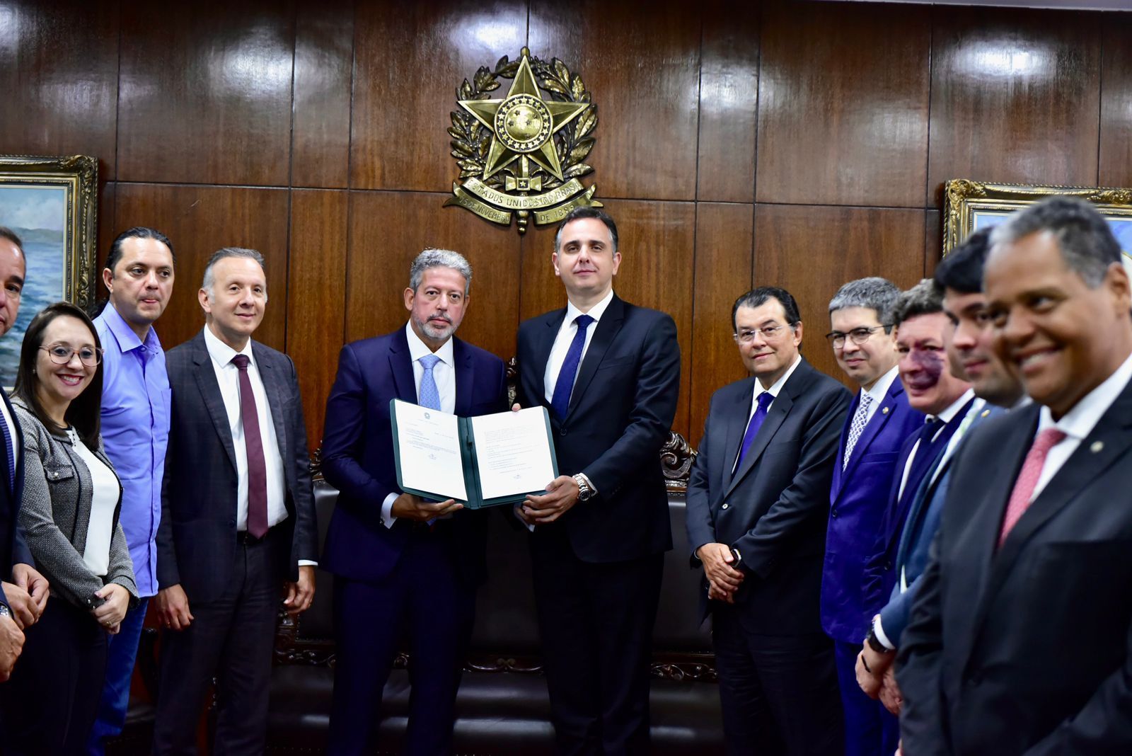 Acompanhado por diversos políticos, o presidente da Câmara, Arthur Lira (à esquerda), entrega uma pasta com o texto da reforma tributária a Rodrigo Pacheco (à direita), presidente do Senado