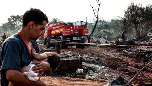 Homem segura criança no colo enquanto bombeiros combatem incêndio em favela