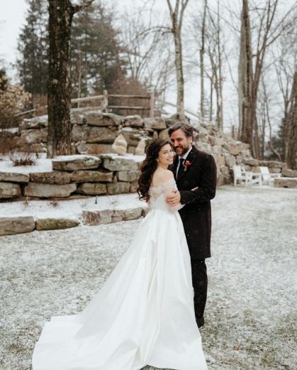 Josh Radnor e Jordana Jacob posam abraçados para foto durante casamento com terno e vestido de noiva em um lugar com pedras ao fundo e muita neve