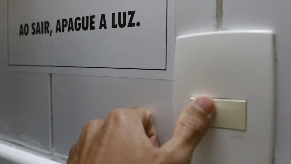 Pessoa desliga interruptor; acima lê-se a frase "ao sair, apague a luz"