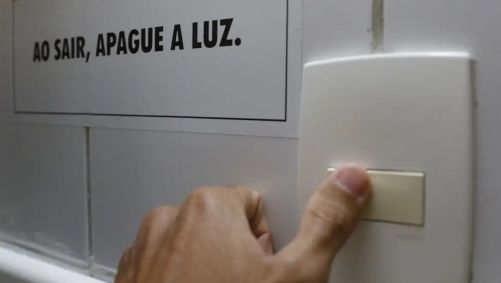 Pessoa desliga interruptor; acima lê-se a frase "ao sair, apague a luz"
