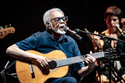 Gilberto Gil. Ele está sentado, cantando num microfone. Ele segura o violão no colo.