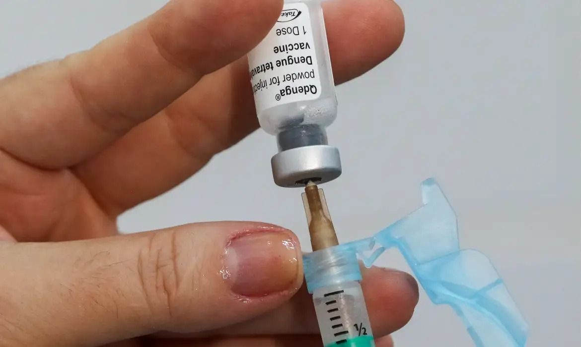 Uma mão segura uma ampola de vacina com uma seringa retirando o líquido