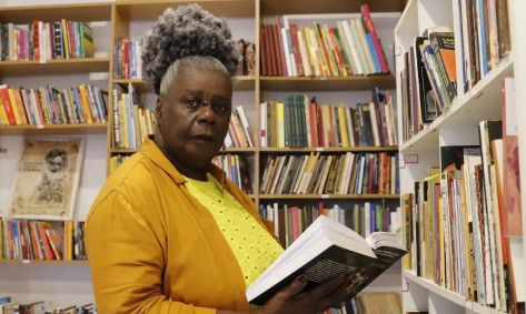 Conceição Evaristo em uma foto na frente de uma estante com livros, segurando um livro aberto nas mãos