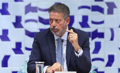 Arthur Lira, sentado, olha para o lado direito enquanto fala e gesticula com a mão esquerda, com o indicador em riste. Na mão direita, segura uma caneta