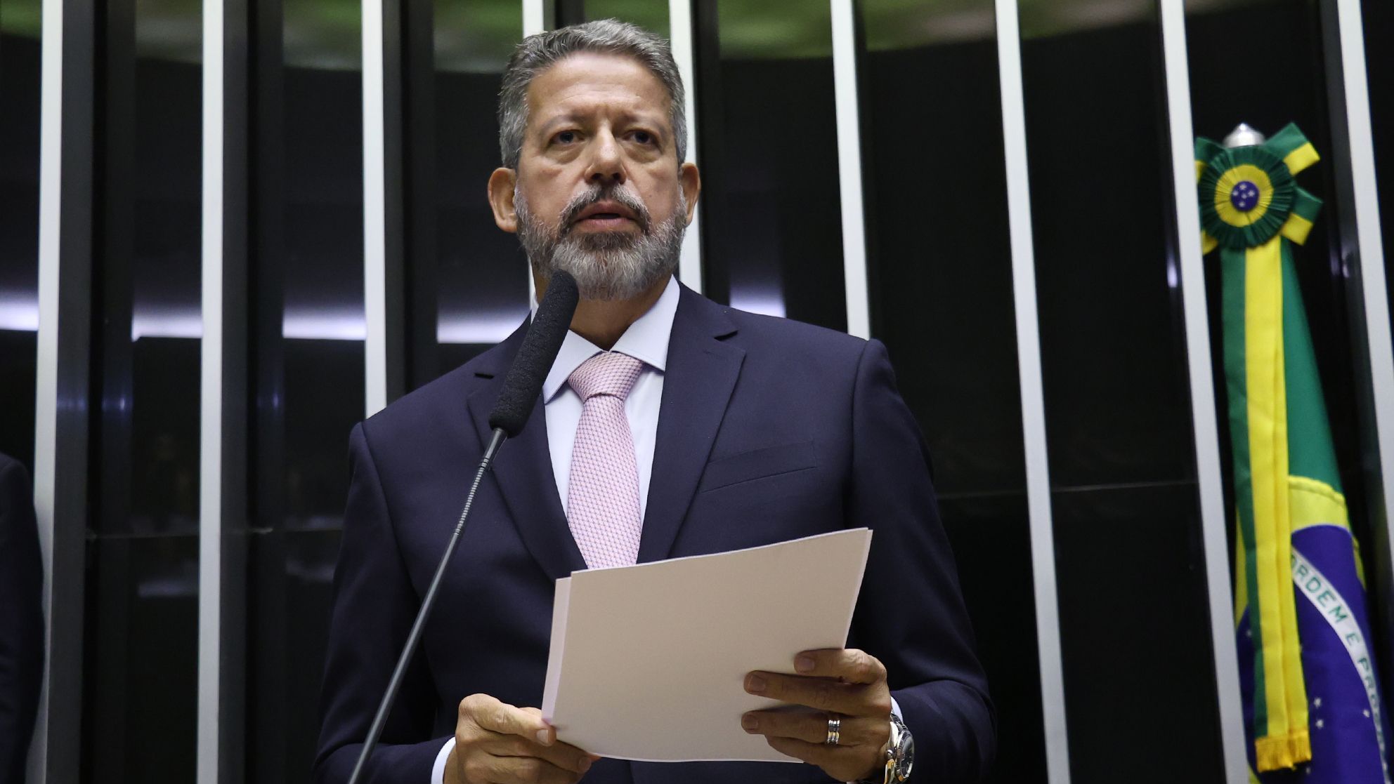 Arthur Lira, de pé diante de microfone, segura papéis nas mãos e olha para frente