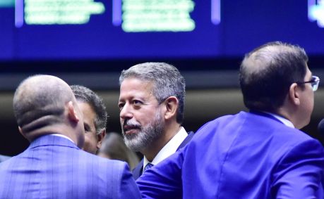 Arthur Lira de perfil com lado esquerdo do rosto em evidência conversa com outro deputado. Outros depois deputados estão de costas, um à esquerda e um à direita de Lira