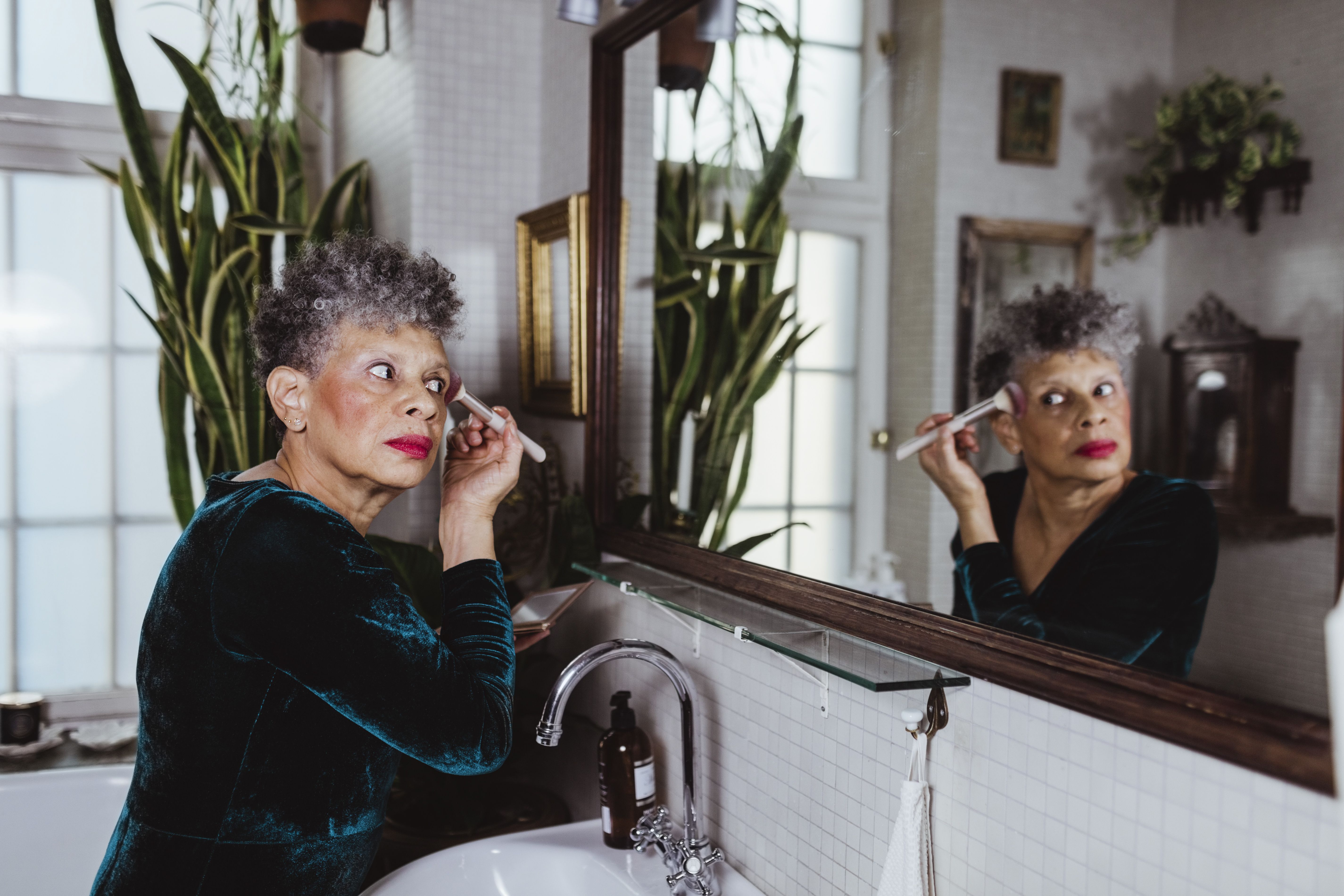 Retired senior female influencer applying make-up while looking at mirror reflection in bathroom