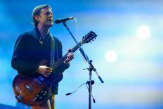 vocalista do king of leon no palco segurando uma guitarra e cantando no microfone com pedestal