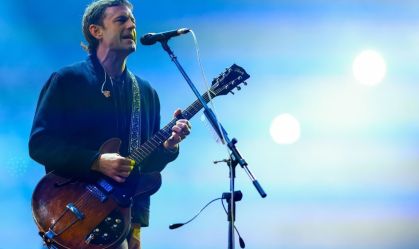 vocalista do king of leon no palco segurando uma guitarra e cantando no microfone com pedestal