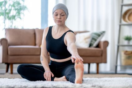 Mulher com lenço na cabeça, veste regata e legging pretas, faz alongamento em sua casa