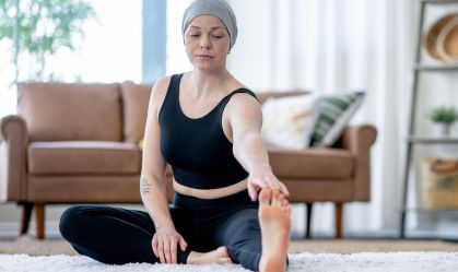Mulher com lenço na cabeça, veste regata e legging pretas, faz alongamento em sua casa