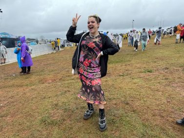 Jovem fazendo o símbolo da paz e com a mão na cintura com várias pessoas de capa de chuva atrás