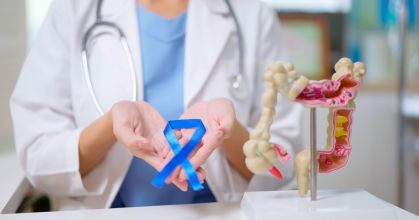 Médica segurando um laço azul, ao lado de uma miniatura de intestino; ao fundo, uma paciente tem seu abdômen examinado por outra médica