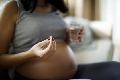 Mulher gestante segurando um comprimido branco em uma mão e um copo d'água em outra