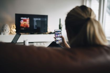 Garota adolescente com cabelos loiros presos em um rabo de cavalo sentada no sofá e assistindo à TV enquanto mexe no celular