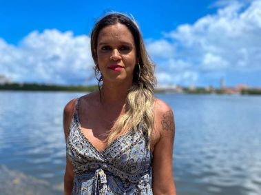A foto mostra uma mulher na frente de um lado. Ela tem cabelos loiros tingidos, usa batom rosa e um vestido estampado azul