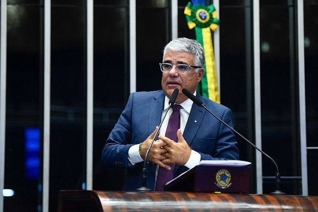 Eduardo Girão veste terno azul escuro e gravata roxa. Tem cabelo branco e usa óculos.