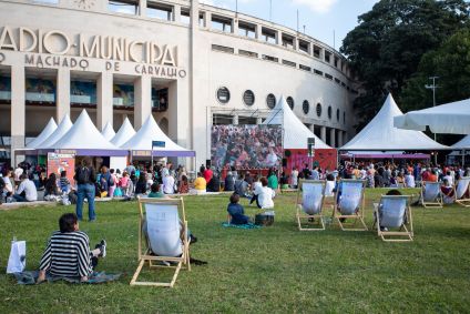 Fachada do Estádio Municipal Paulo Machado de Carvalho. Há tendas de livros e pessoas no local. O ambiente é aberto.