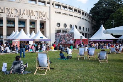 Fachada do Estádio Municipal Paulo Machado de Carvalho. Há tendas de livros e pessoas no local. O ambiente é aberto.