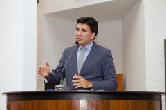 Júnior Geo veste terno cinza, camisa azul e gravata cinza. É branco e tem cabelo preto.