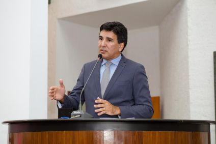 Júnior Geo veste terno cinza, camisa azul e gravata cinza. É branco e tem cabelo preto.
