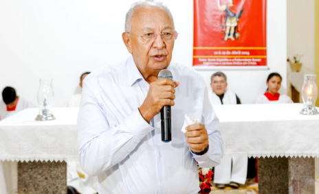 Foto mostra Dr. Pessoa segurando o microfone em uma missa. Ele é idoso, usa óculos e usa uma camiseta branca.