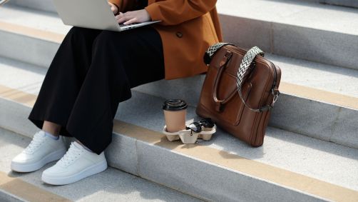 pessoa sentada em escada digitando num laptop. Ao seu lado, um copo de café e uma pasta