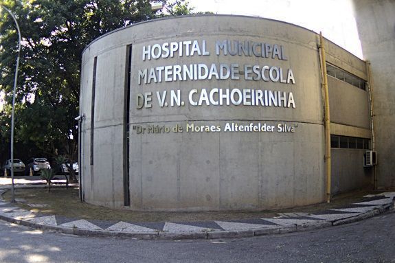 Foto mostra a fachada do Hospital Municipal Maternidade Escola de V.N. Cachoeirinha, em São Paulo. A fachada é inza, de concreto, e o nome do hospital está em alto relevo, prateado.