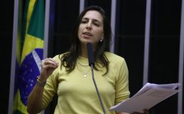 Natália Bonavides veste camisa de manga amarela, colares e relógio. É branca e tem cabelo médio castanho escuro.