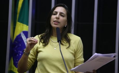 Natália Bonavides veste camisa de manga amarela, colares e relógio. É branca e tem cabelo médio castanho escuro.