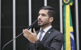 André Fernandes é branco, tem barba e cabelos castanhos, veste terno preto, gravata cinza e camiseta branca