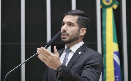André Fernandes é branco, tem barba e cabelos castanhos, veste terno preto, gravata cinza e camiseta branca