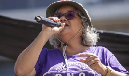 Lenilda Luna veste camisa roxa, chapéu de tecido, óculos escuros e segura um microfone