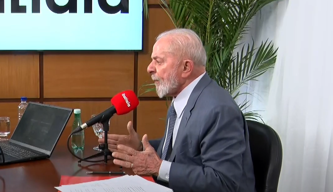 Foto mostra presidente Lula. Ele veste um terno cinza, camiseta branca e gravata. É idoso, tem barba branca. Ele fala com um microfone vermelho à frente.